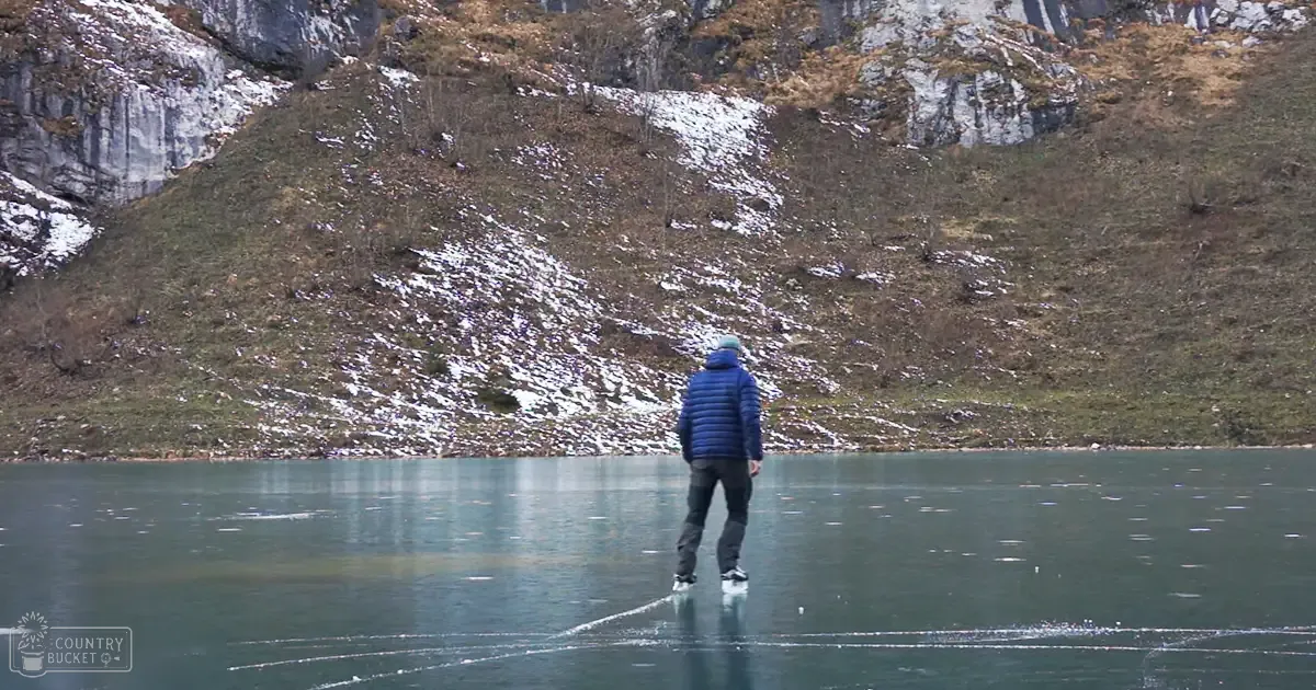 Adult male in dark blue puffer jacket, black pants, white shoes on icy frozen lakeshore with mountainous terrain and diffused winter light.