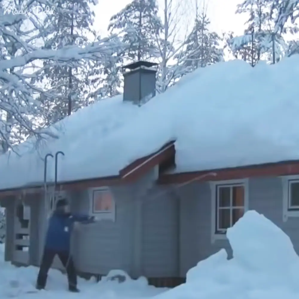 An adult male shoveling snow near a house in a snowy residential area with trees and bright daylight, wearing a dark blue jacket with long sleeves and thick-soled black boots.