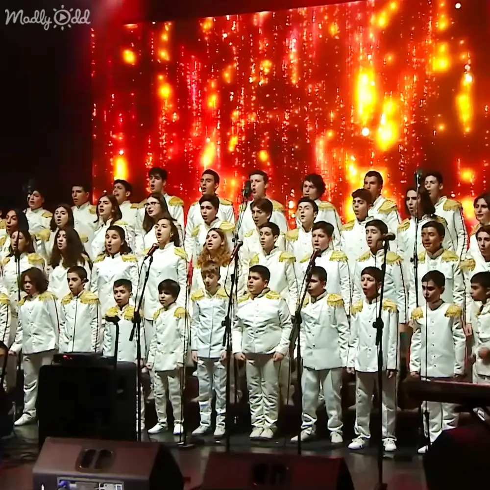 A choir of young male and female performers in white uniforms with gold trimmings, performing on a stage illuminated by red lighting effects resembling falling lights or fireworks.
