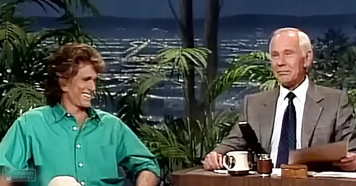 A young adult male in a bright green button-up shirt sits at a table with an older adult male dressed in a dark suit, on what appears to be a talk show set with decorative plants and scenic outdoor views.