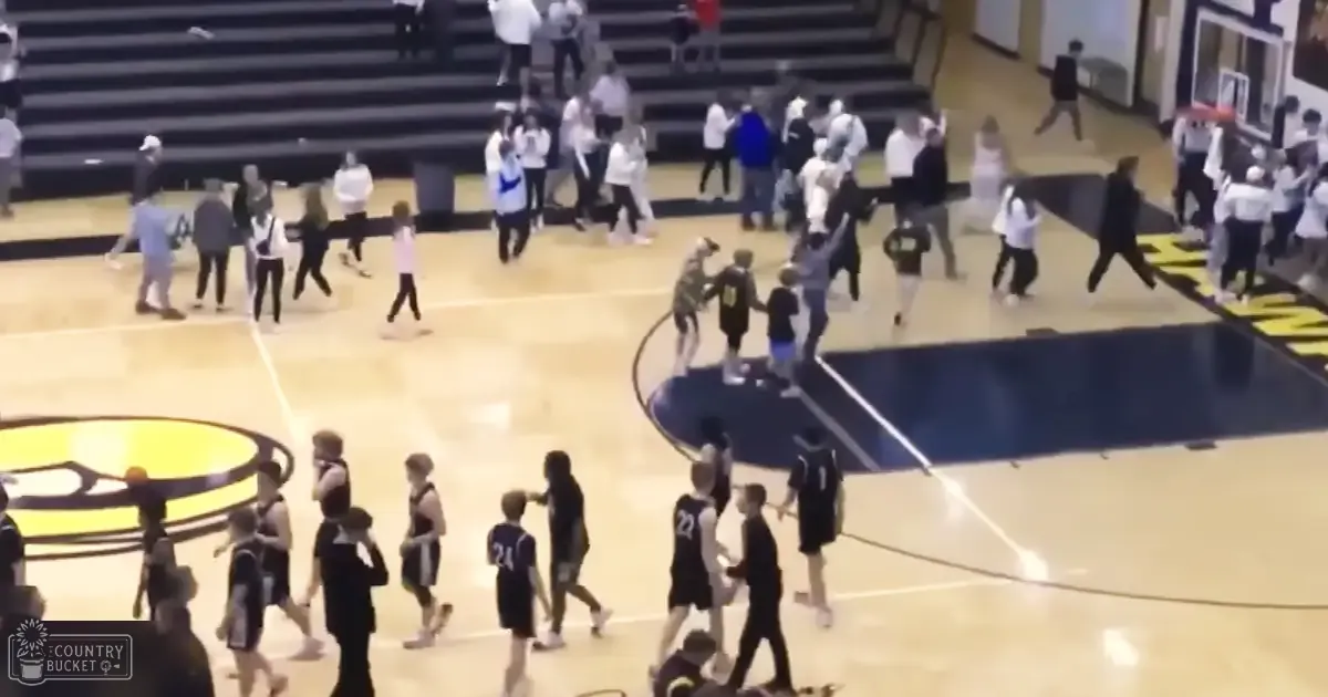 Youth basketball game in progress at an indoor sports facility; players in dark jerseys and athletic shorts, spectators seated on bleachers under artificial lighting.