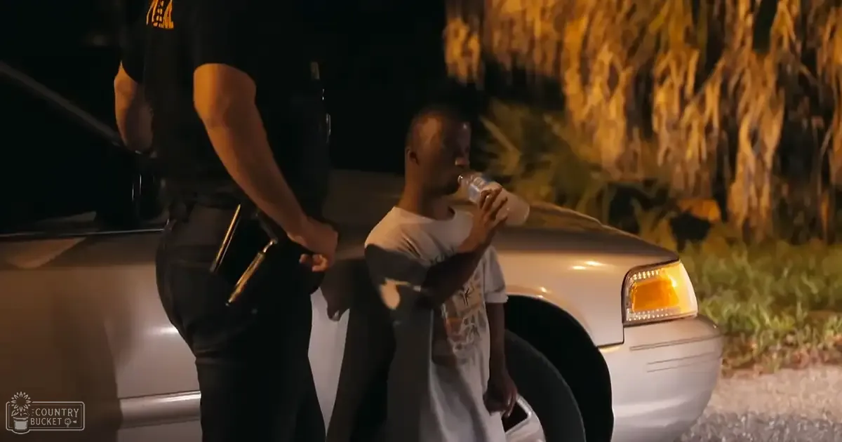 An adult male police officer in a dark uniform interacting with a young adult to middle-aged man wearing a casual white t-shirt near a white sedan at night, illuminated by artificial light and trees or bushes in the background.