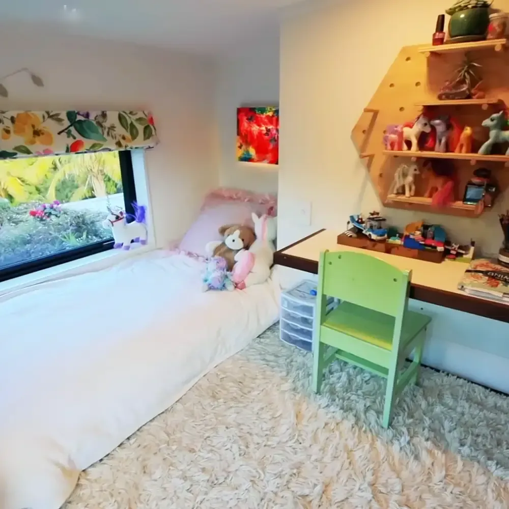 Child's bedroom with white bedding, green chair, floral curtains, colorful abstract painting, toys and books on shelves, indoor setting in a home environment during daytime.