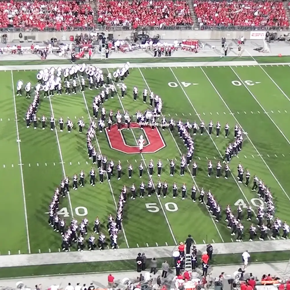 A marching band performing at a football stadium with spectators in red attire during daylight under clear skies.