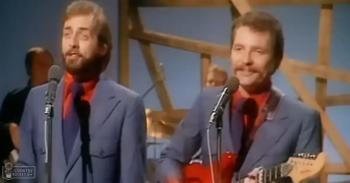 A musical performance scene with two adult male performers in light blue suits and red shirts, set on a stage with microphones and wooden scaffolding under bright even lighting.