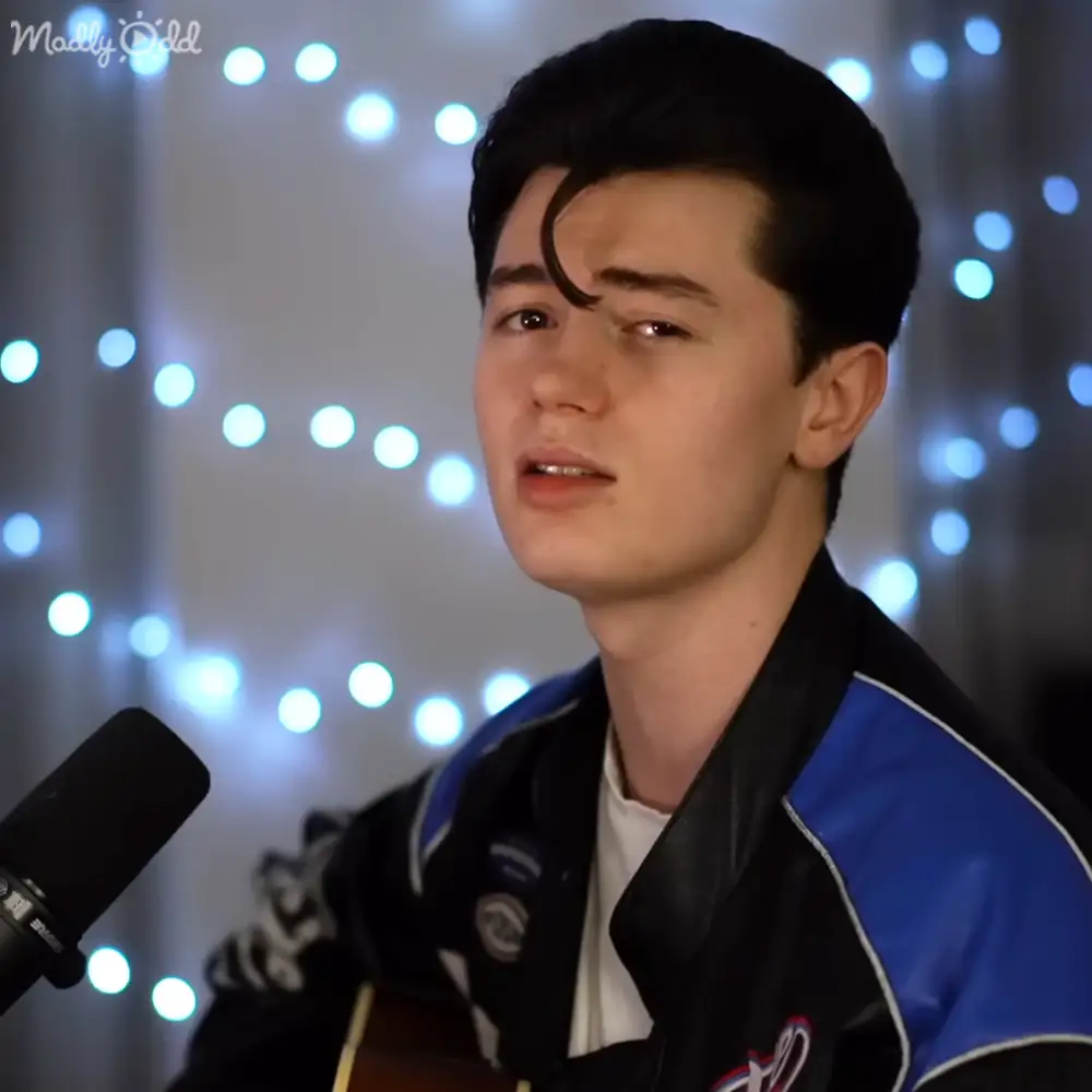 Young adult male wearing a black and blue synthetic jacket with high collar, standing indoors near a microphone and string-lit backdrop.