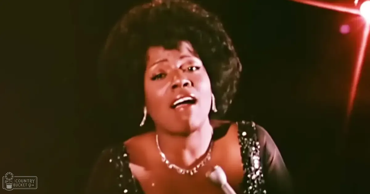 An adult female performer in a 1970s or early 1980s black sequined dress, holding a microphone on stage under red lighting.