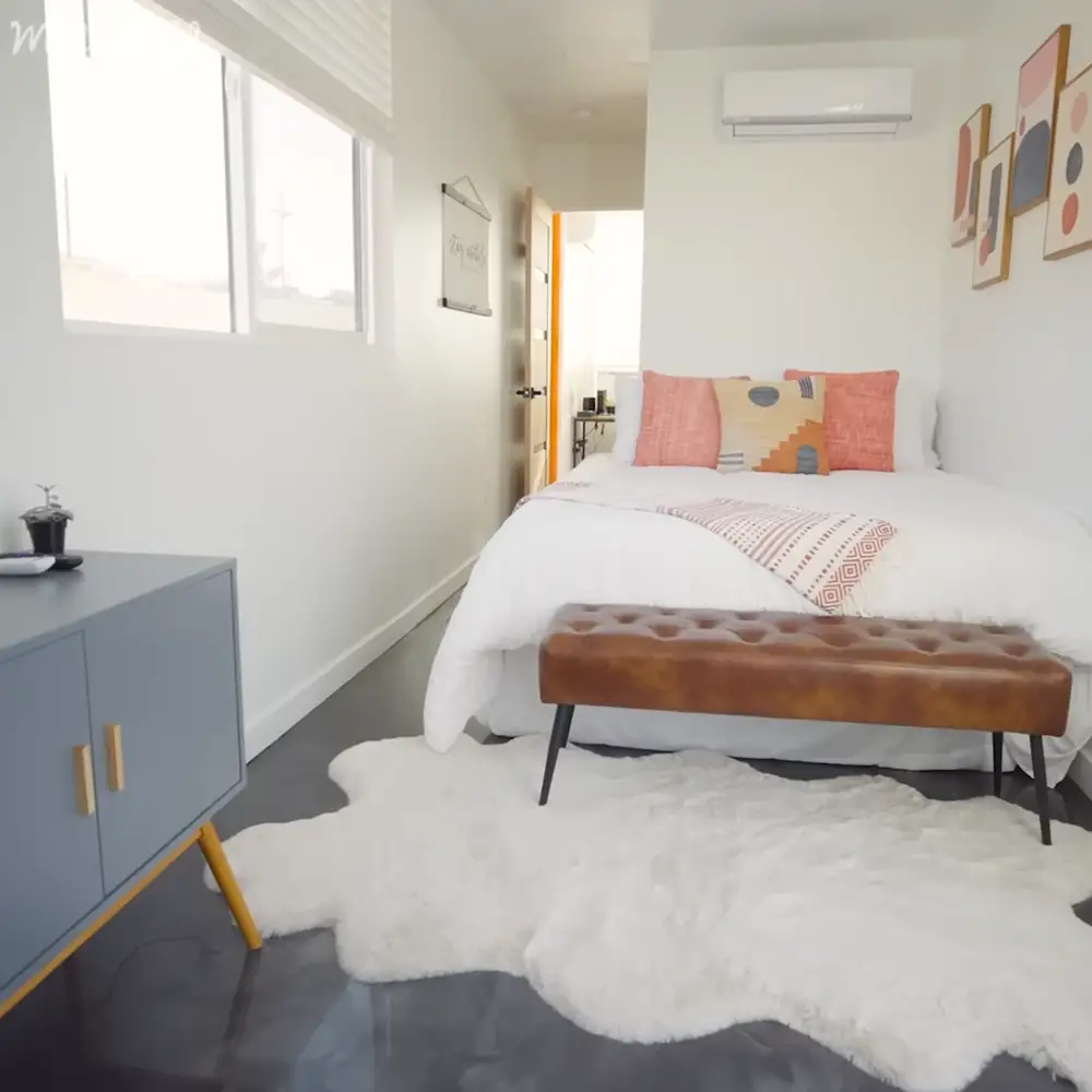 A cozy bedroom scene with a bed with white bedding, orange and pink pillows, and a patterned throw; brown leather tufted bench at the foot of the bed; blue cabinet with gold handles on the left side; white fluffy rug under the bed; wall art with abstract designs in red, black, and beige tones; small desk or table near the window with a plant on it.