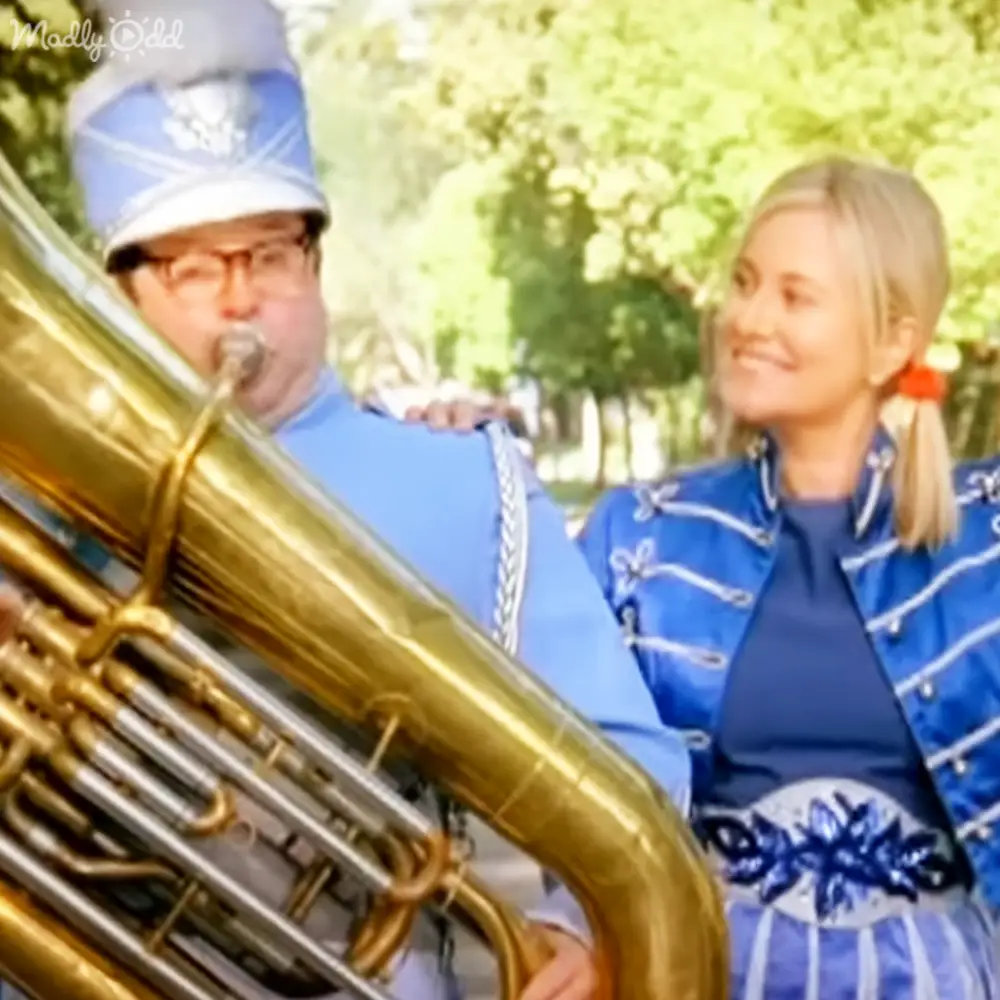 A middle-aged male tuba player in a blue marching band uniform stands outdoors with a young adult female wearing a striped jacket, amidst greenery and natural daylight.
