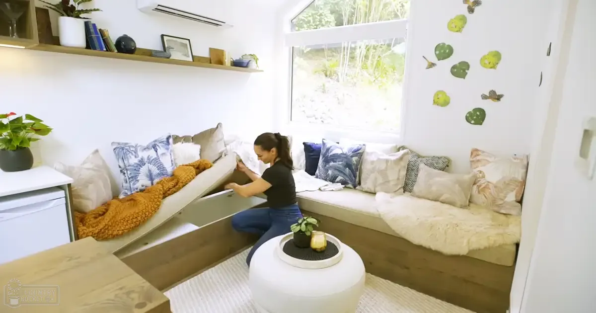 A woman in a living room with large windows and greenery outside, wearing a black t-shirt and blue jeans.