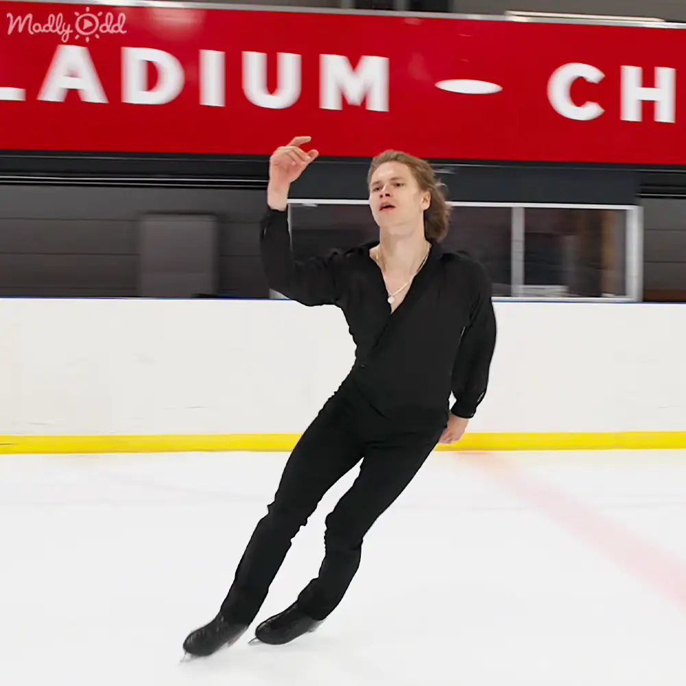 A young adult male figure skating in an indoor ice rink with a red sign reading "ADIUM - CH" above him, wearing a black synthetic v-neck outfit and a necklace.