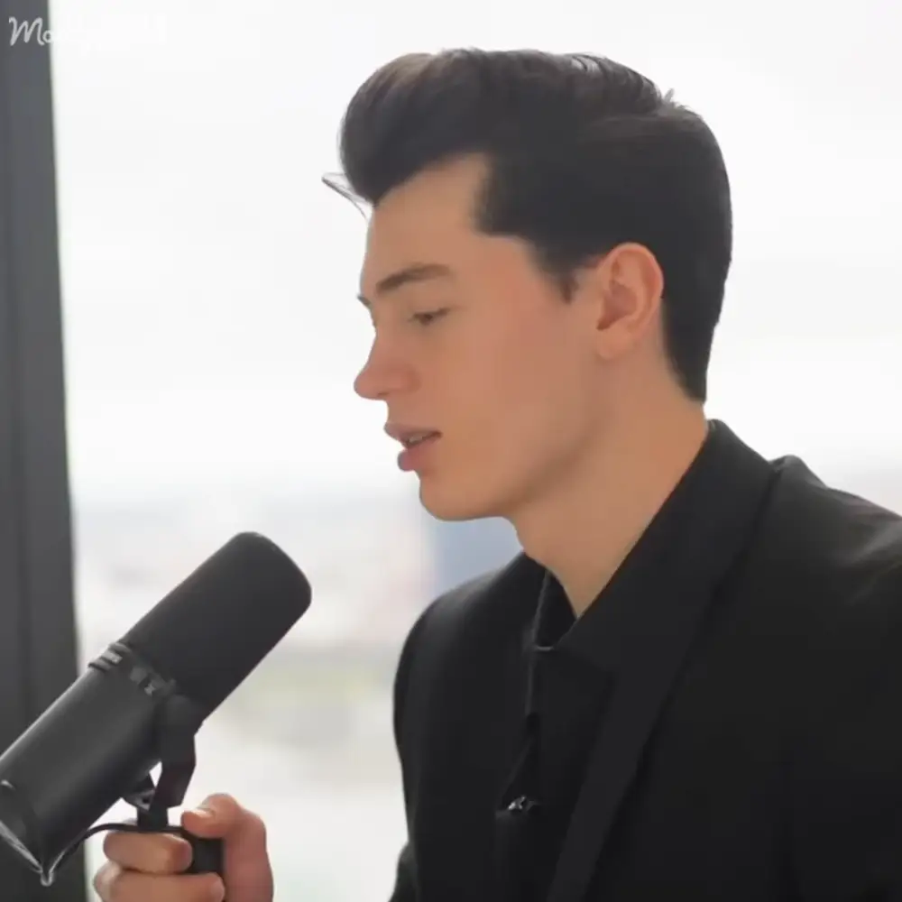 A young adult male in a tailored black suit jacket stands indoors, holding a professional condenser microphone with a pop filter attached.