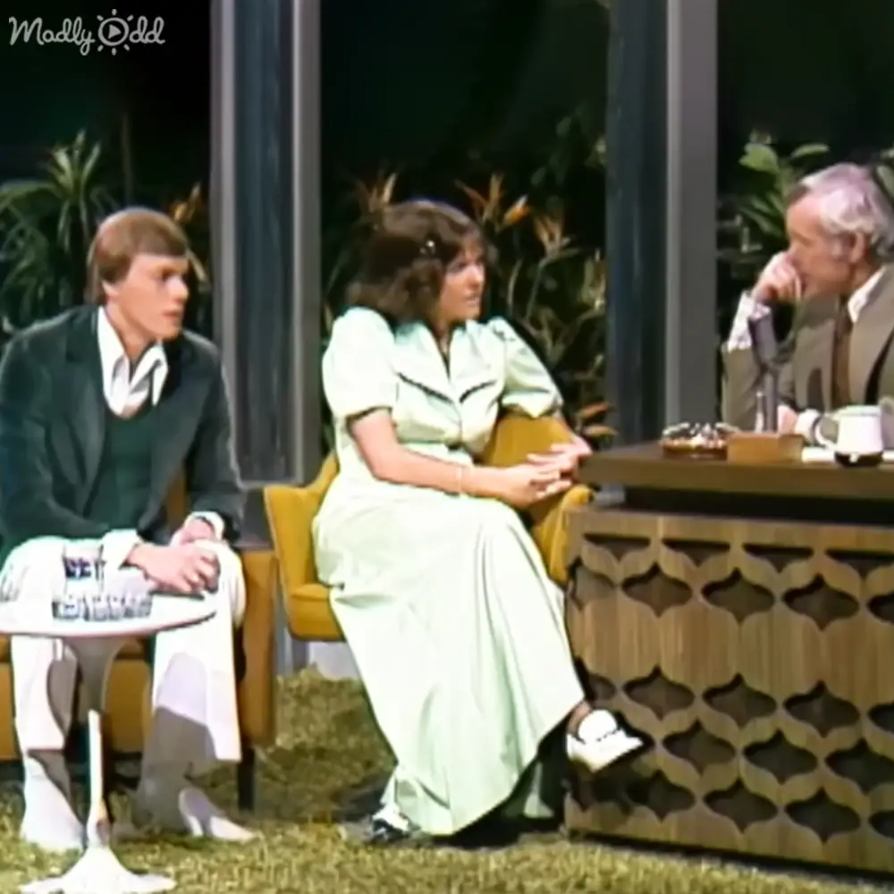 A woman in an adult years' dress seated between two men, likely part of an interview or talk show set.
