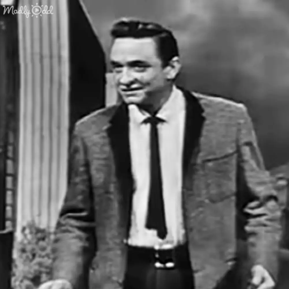 Young adult male wearing a textured blazer and tie, standing in an indoor setting with stage lighting and dark backdrop.