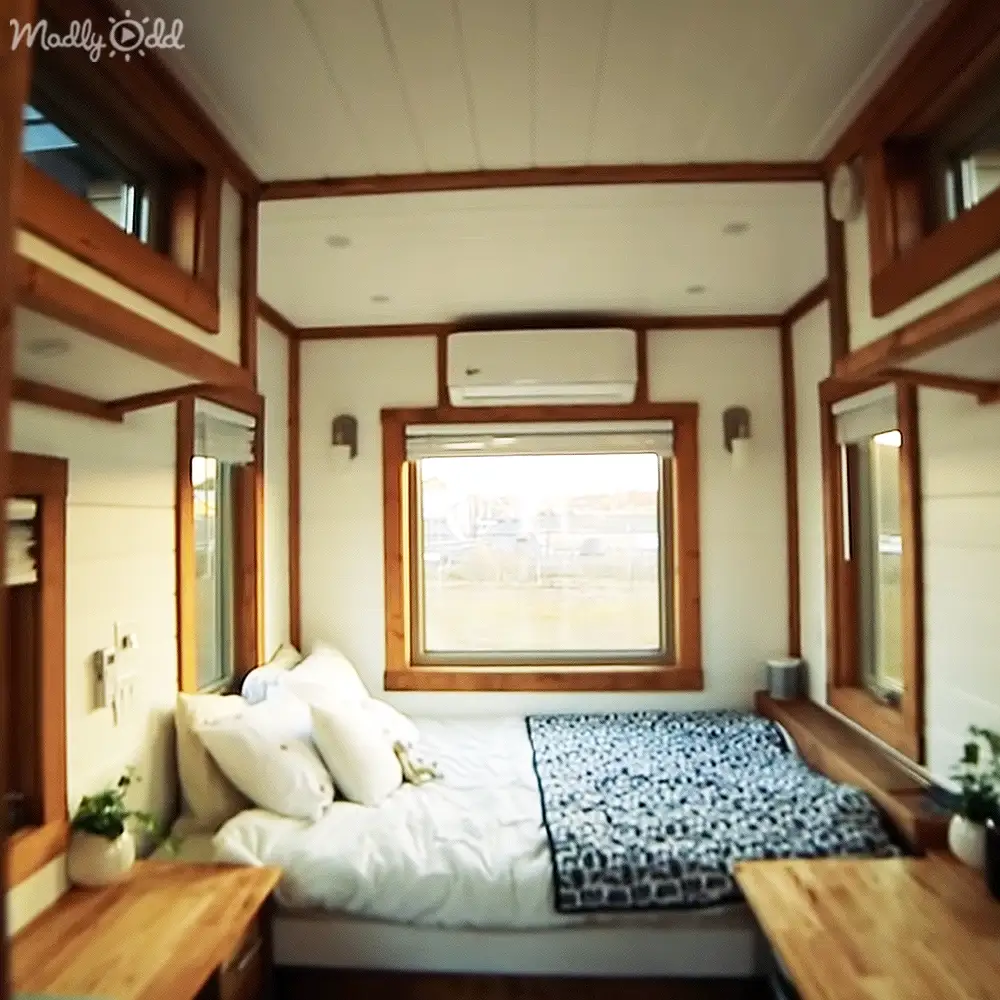 Cozy bedroom setup with white bedding, black patterned throw blanket, wooden desks, window air conditioner, small potted plants, wall sconces in a small house or cabin during daytime.