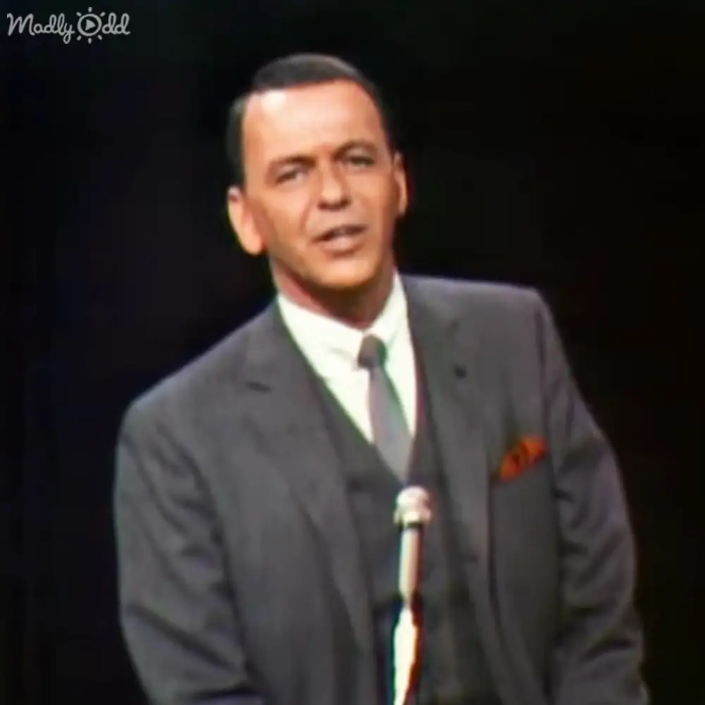 An adult male performer in a dark gray suit with a white shirt and dark tie, holding an orange or red pocket square on stage under bright lighting.