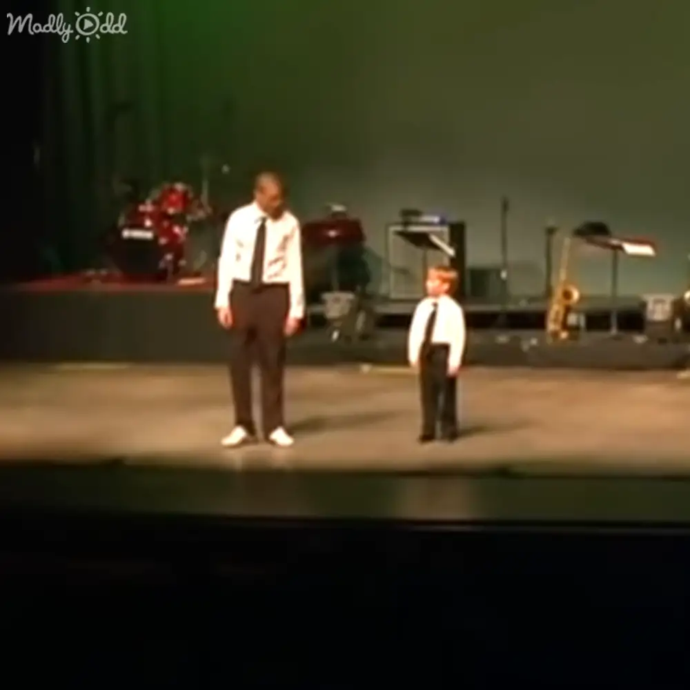 A middle-aged man and a toddler wearing matching white shirts with dark ties stand together on a dimly lit stage surrounded by musical instruments.