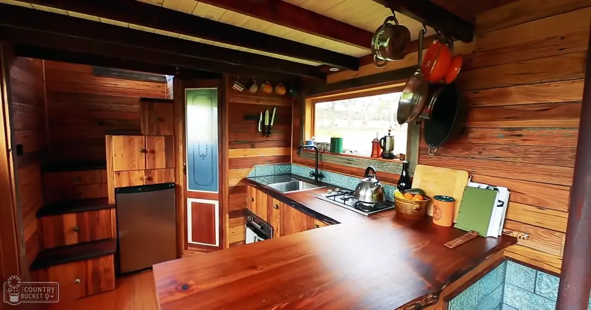 A cozy kitchen setup with wooden cabinets, hanging pots, a magnetic knife strip, green cutting board, orange basket with fruits, black kettle, and other kitchen utensils.