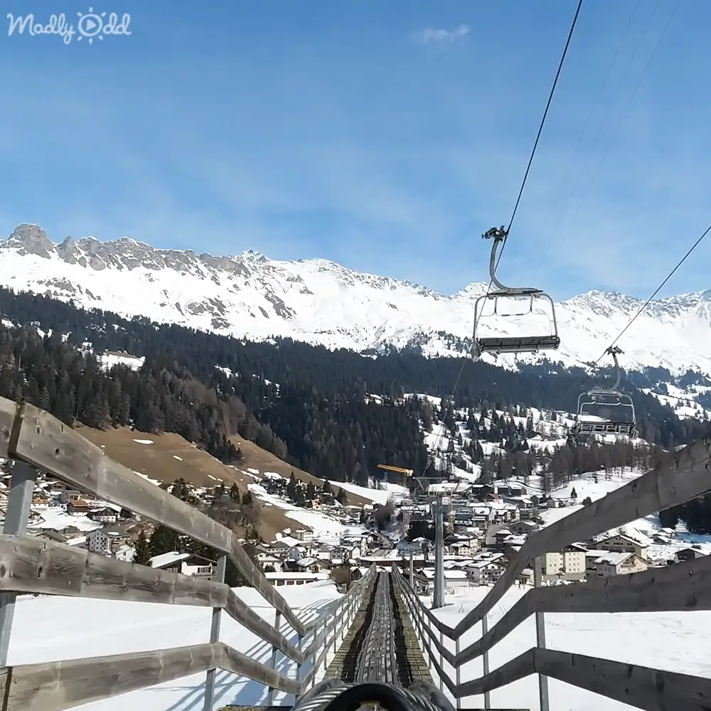 A ski lift with wooden railings, leading down towards a village at the base of snow-covered mountains and dense evergreen forests under bright daylight.