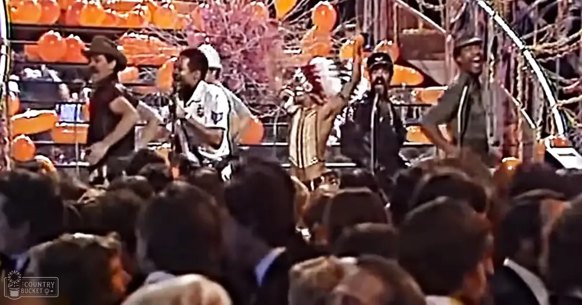 An indoor concert scene with performers wearing dark and patterned outfits under artificial stage lighting, with orange pumpkins on a backdrop suggesting an autumn theme.