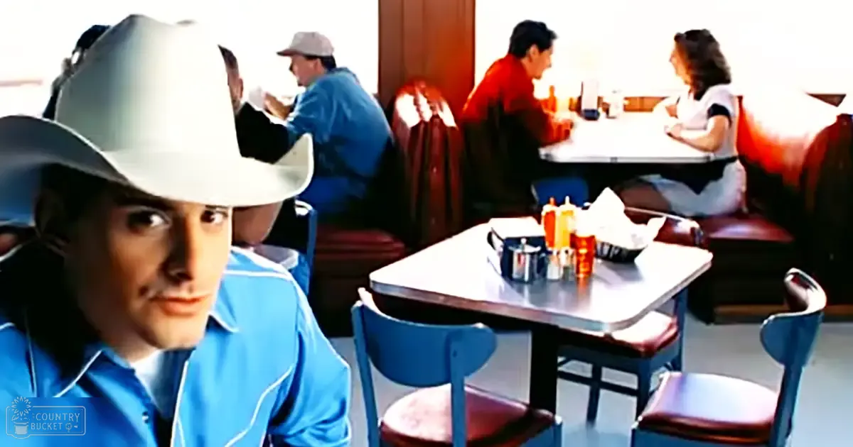 A young adult male wearing a cowboy hat in a diner setting with other patrons seated at tables, condiments on the table, and red cushioned chairs.
