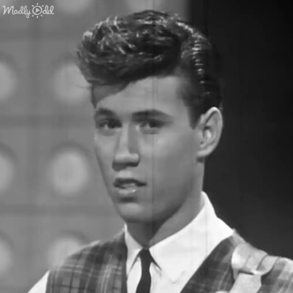 Young adult male wearing a collared white undershirt with a plaid shirt over it, standing against a patterned wall background under artificial studio lighting.