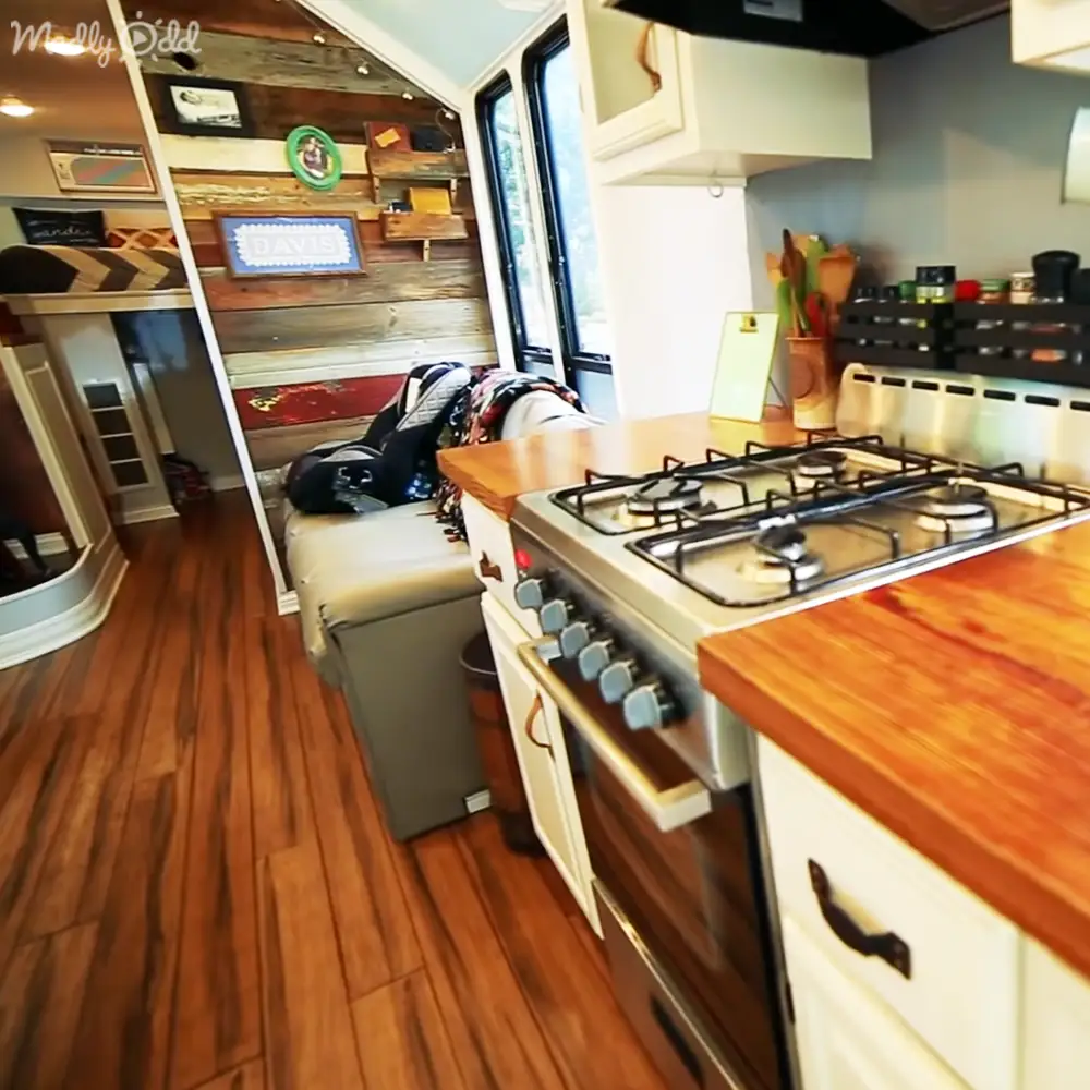Compact kitchen area within a mobile home or RV, with gas stove, wooden countertop, white cabinets, and decorative items like framed pictures and signs.
