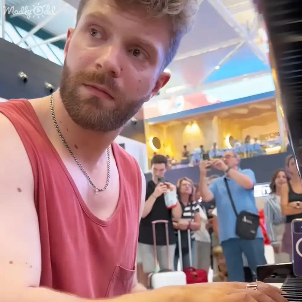An adult male singer in a red sleeveless tank top seated at an indoor public space with high ceilings and natural light, possibly an airport or shopping mall.