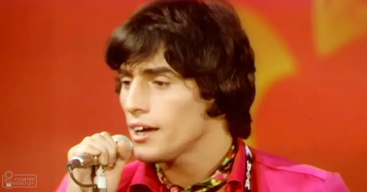 A young adult male in a relaxed, brightly colored outfit singing into a microphone on an indoor stage with a vibrant backdrop.