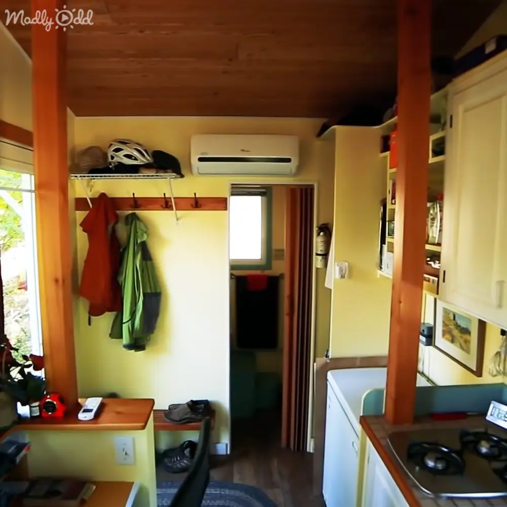 A cozy interior space with a wooden ceiling, wall-mounted coat rack holding jackets and a helmet, white air conditioning unit above the rack, small table with red clock and white phone, kitchen area with cabinets, stove, sink; natural light through window; yellow walls, white appliances, brown ceiling, black jackets/shoes, green jacket, red clock, gray sink