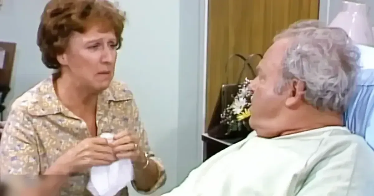 Middle-aged female wearing a floral print blouse in a hospital room, lying beside an older adult male in light green gown.