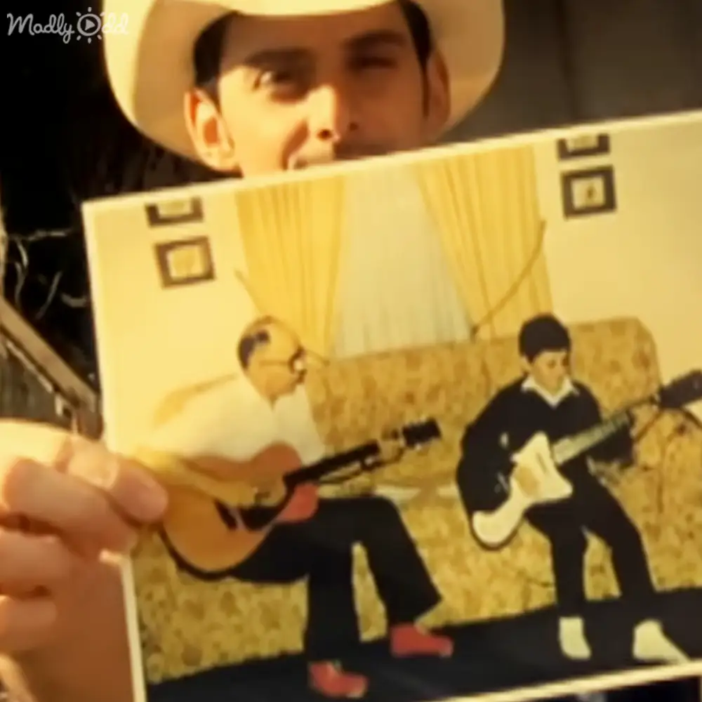 A young adult male holds an album cover with two guitarists in a retro-style setting. The scene is indoors with soft evening lighting, and the person wears a cowboy hat.
