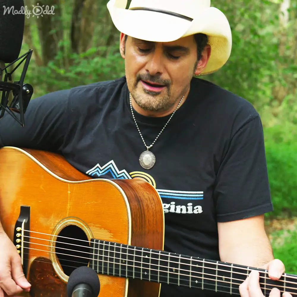A middle-aged adult male wearing a black 'Virginia' t-shirt with decorative lines, playing an acoustic guitar in an outdoor setting with lush greenery, and a microphone positioned nearby for recording or performance.
