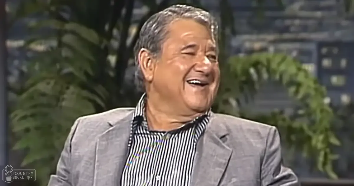 An older adult male wearing a gray suit jacket over a black and white striped shirt, set against an indoor studio backdrop with a cityscape view.