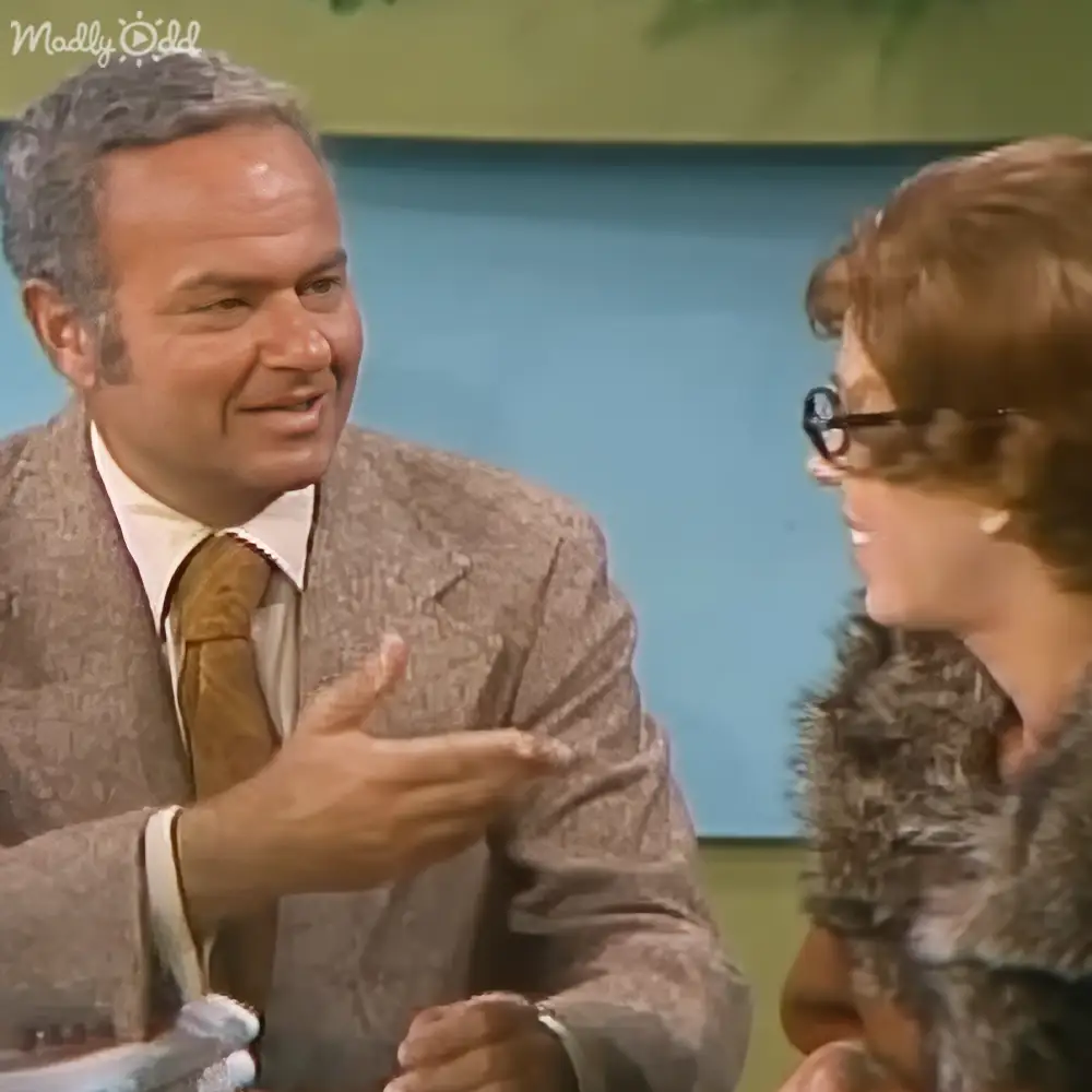 Adult male wearing a patterned suit jacket and brown tie indoors with an adult female in a dark-colored dress holding black-rimmed glasses nearby.