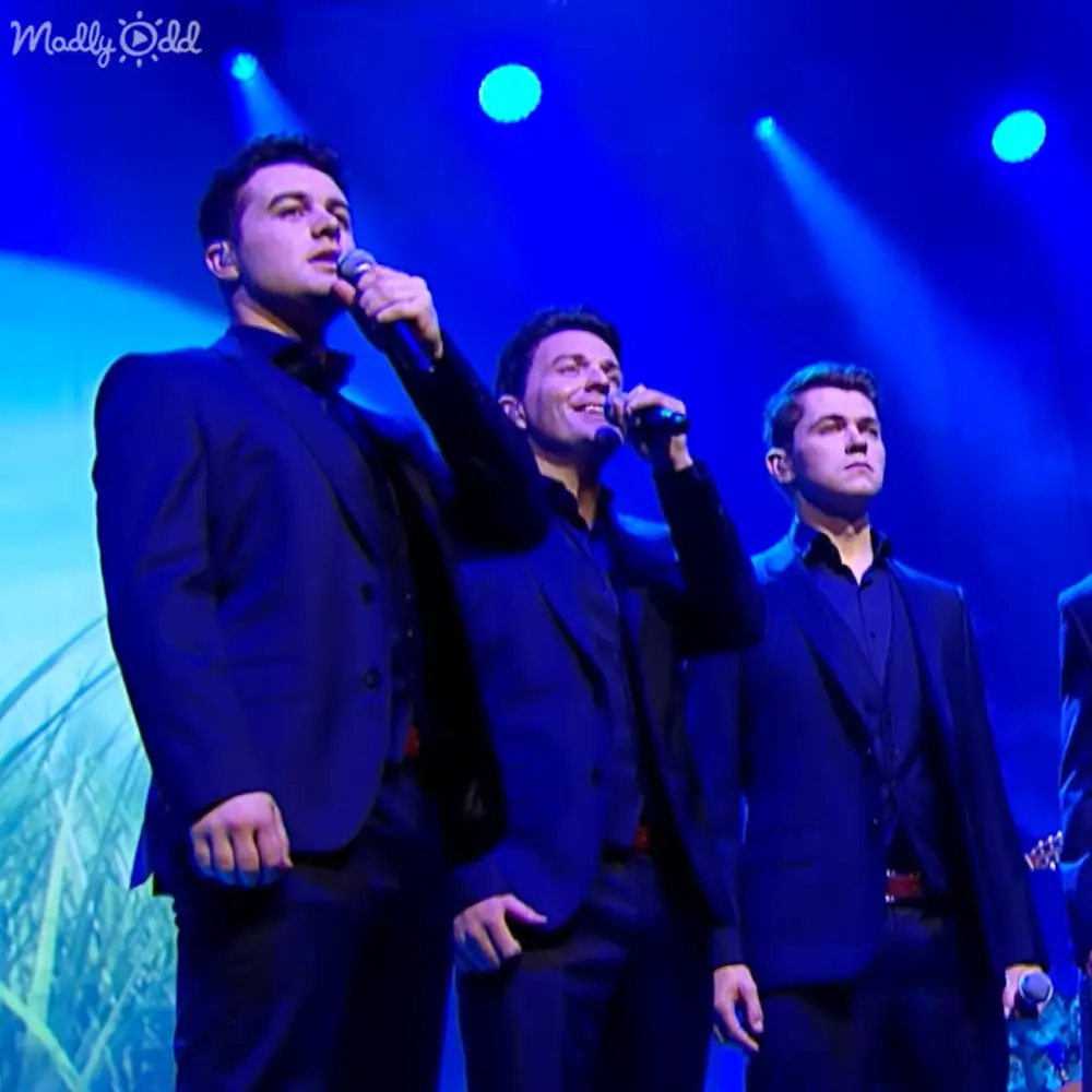 An indoor musical performance scene with three adult males in dark suits, blue lighting and a grassy image backdrop; black suits against blue stage lights