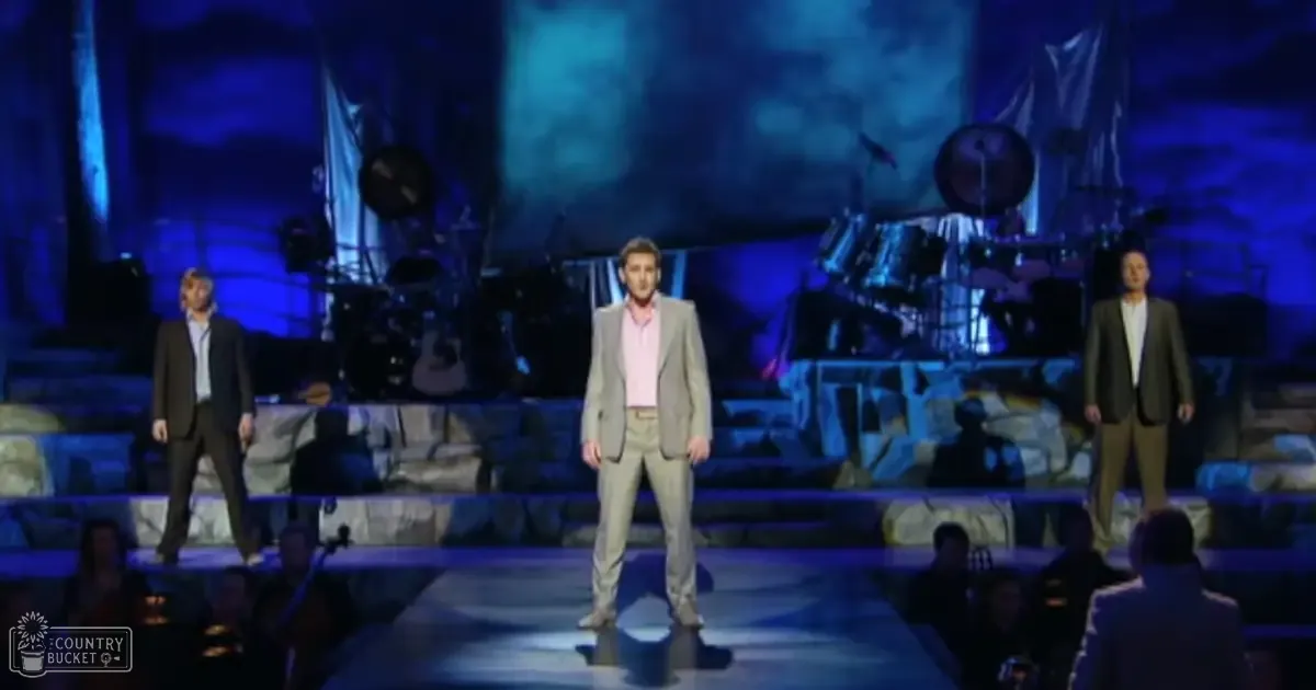Performers in a middle-aged adult male singer wearing a light beige suit and pink shirt, surrounded by young to adult males in dark suits on an indoor concert stage with abstract blue visuals.