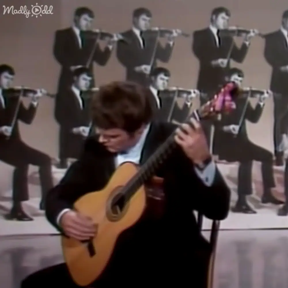 A young adult male guitarist in a black suit jacket, white shirt with a black tie, and dark pants plays guitar against a backdrop of multiple images of men playing flutes. The setting suggests an indoor performance or recording studio environment.