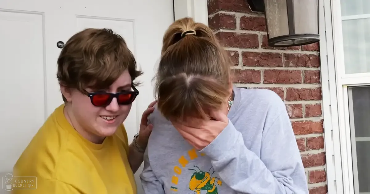 A young adult female in a yellow T-shirt stands outside near a brick wall, wearing red-tinted sunglasses with a white door and light fixture behind her.