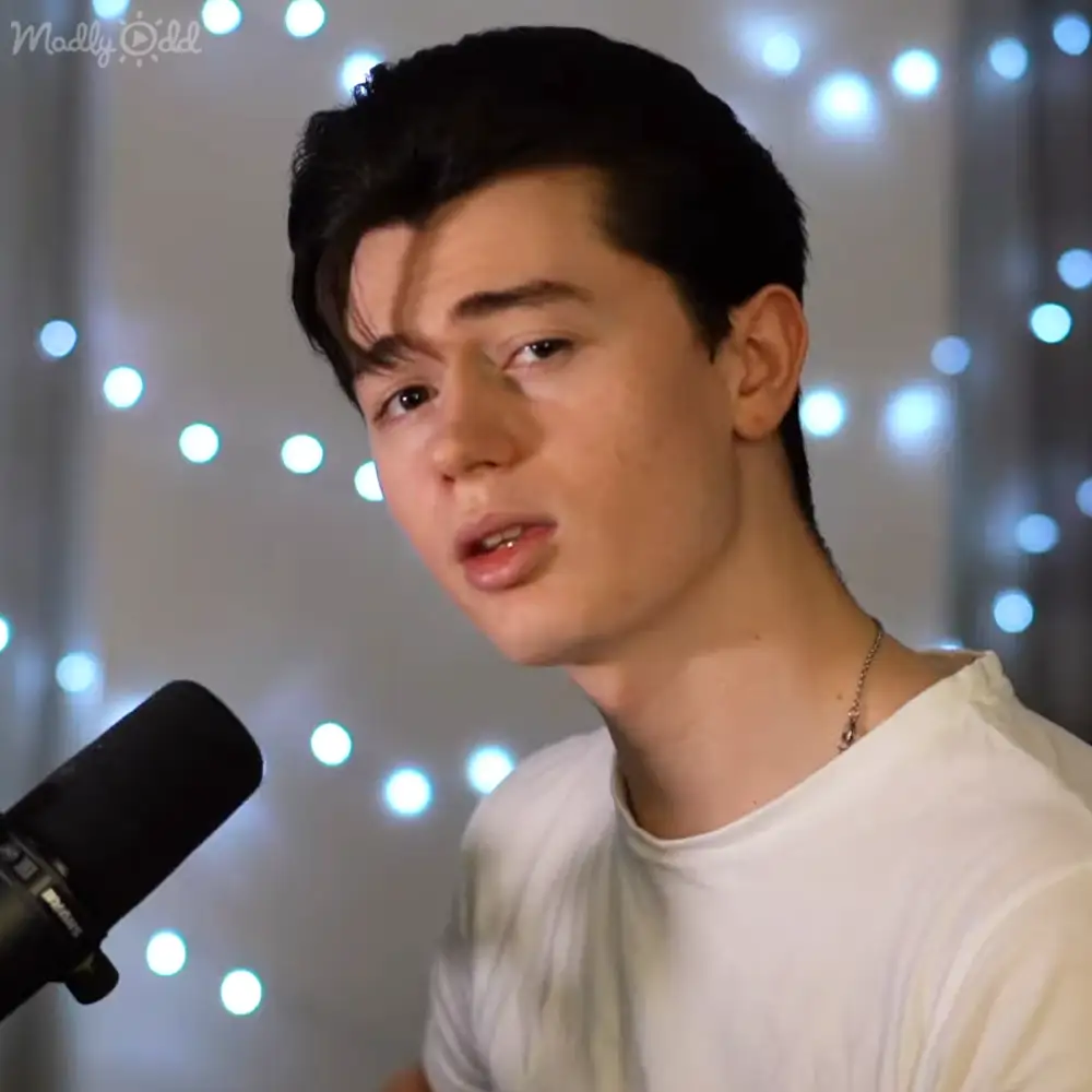 A young adult male in a casual white t-shirt with string lights and a microphone, suggesting an indoor recording or streaming setup.