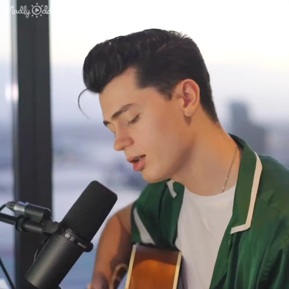Young adult male in a relaxed green jacket with white t-shirt, holding a guitar indoors near a cityscape view.