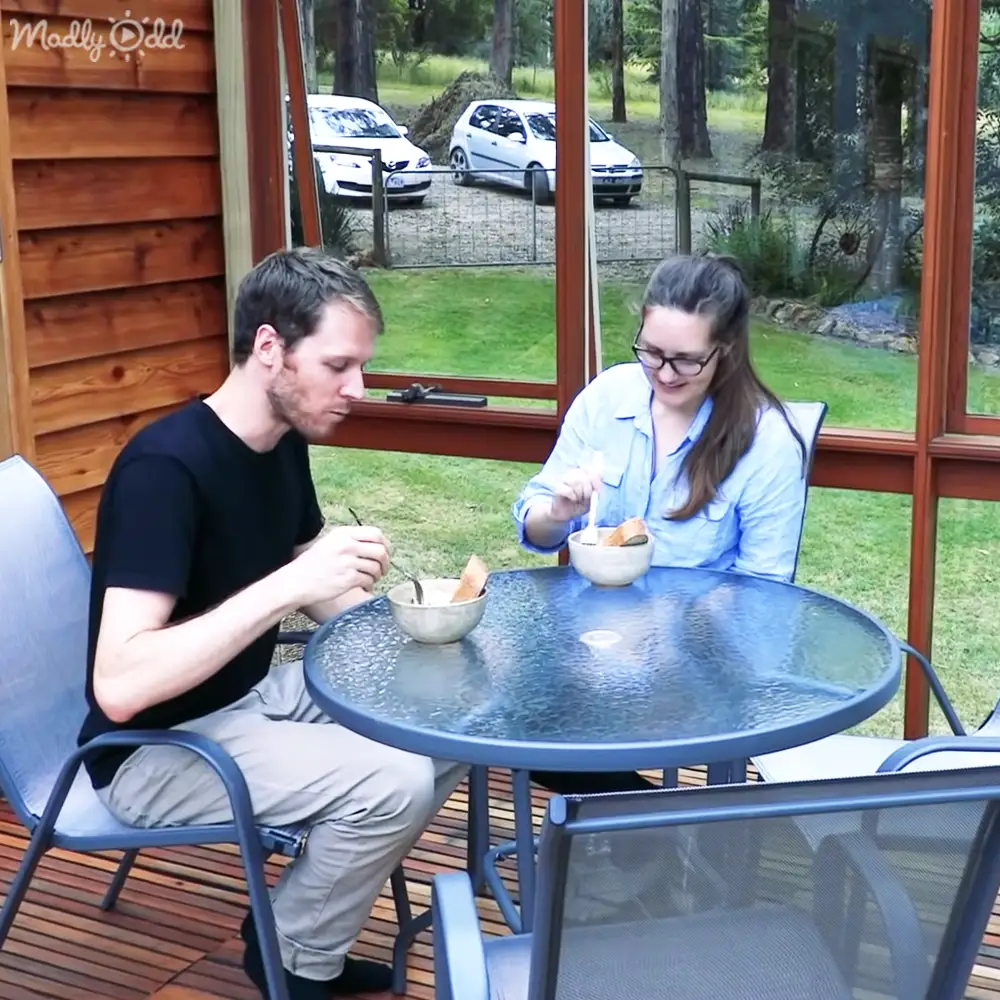 A young adult male and an adult female enjoying a meal outdoors on a patio surrounded by greenery, with natural daylight illuminating the scene.