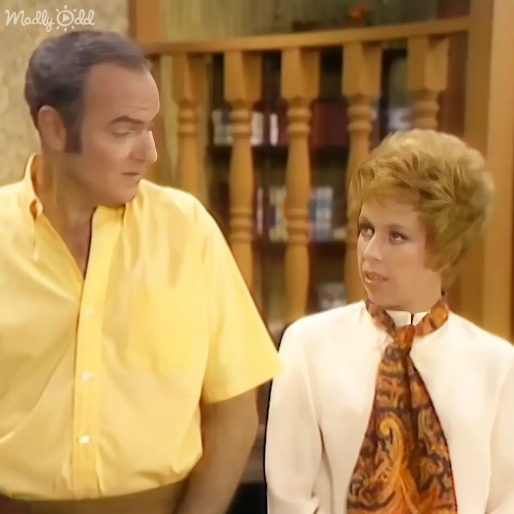 A child in a white blazer over an orange and brown patterned tie stands next to an adult male in a bright yellow shirt, both indoors with warm lighting and wooden staircase railing visible.