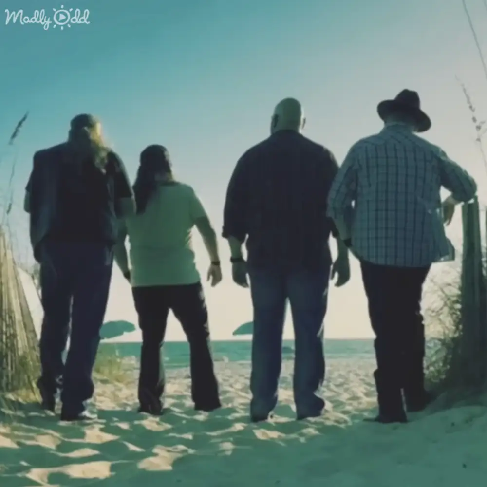 Four individuals standing together at a beach; older adult male wearing a dark suit jacket and trousers, adult female in a light green t-shirt, middle-aged male in a dark jacket and jeans, older adult male with a plaid shirt and black hat;
