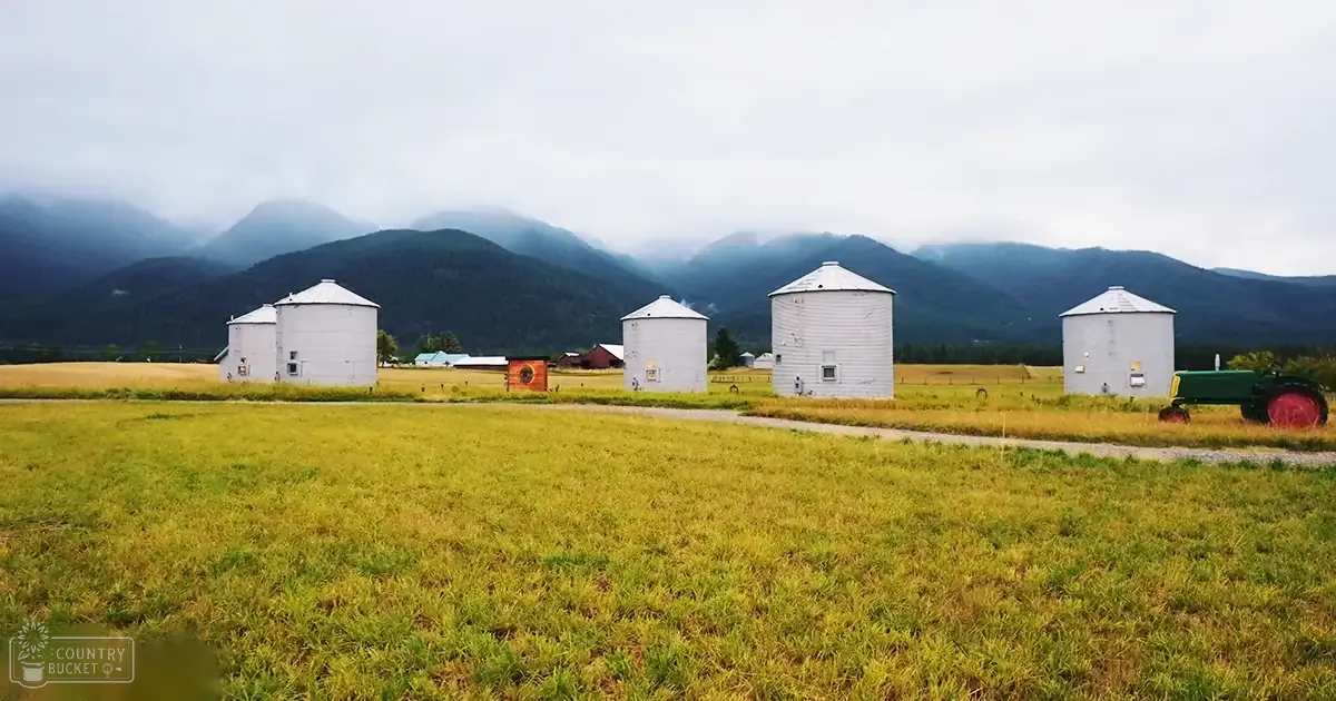 A serene rural farm scene with white cylindrical silos, a green tractor with red wheels, and mountains partially covered by mist in the background. The setting is likely early morning or late afternoon.