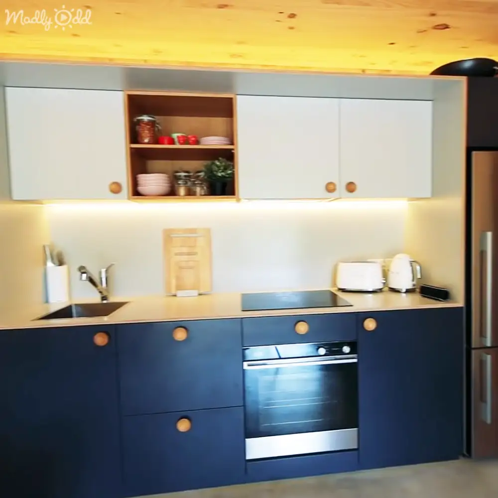 Modern kitchen with blue and white cabinetry, wooden shelves holding dishes and jars, sink, knives block, cutting board, toaster, kettle, oven; indoors residential home setting, warm lighting suggests daytime or early evening.