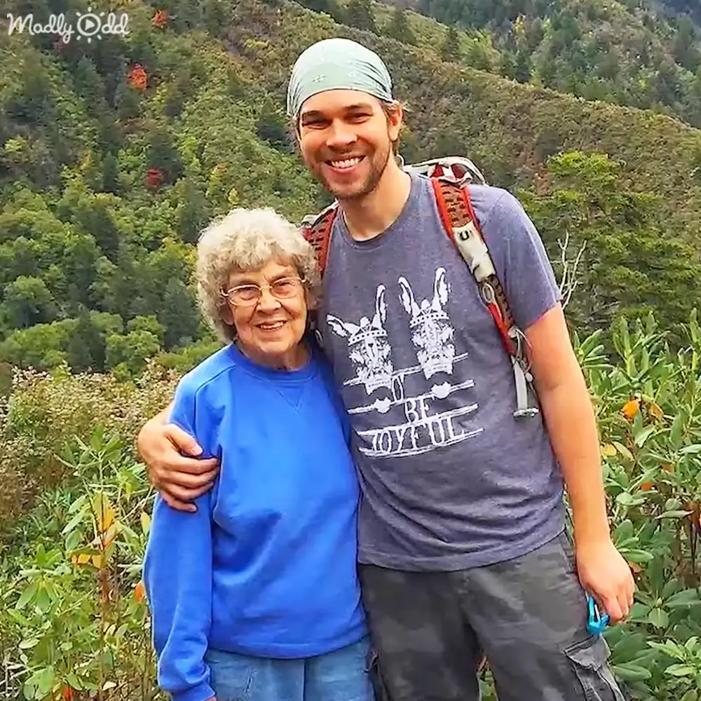 An older adult female with short curly gray hair, glasses, blue shirt and light-colored pants, standing outdoors in a lush green forested area. A young adult male with short hair wearing a light blue bandana, dark gray t-shirt with 'BE JOYFUL' text, black cargo pants, red backpack strap, and small object in hand is seen alongside her.