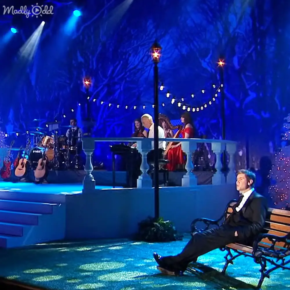 An adult male wearing a dark suit with a white shirt and tie sits on a bench in an outdoor stage setting at night, surrounded by string lights and musical instruments.