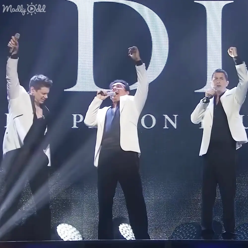 Three adult male performers on stage, likely at a concert or event, illuminated by bright lighting with a dark background and a large screen displaying bold text in the backdrop.