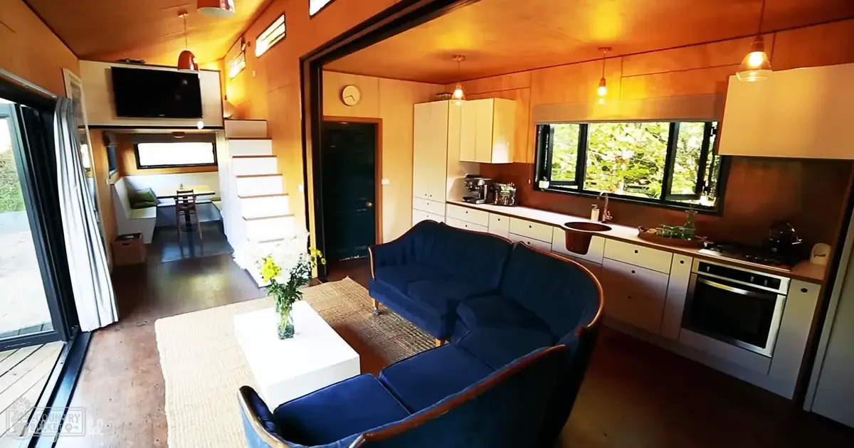 Compact modern living space with a blue curved sofa, white coffee table with flowers, and small dining area with chairs; indoor residential setting with natural light and greenery outside.
